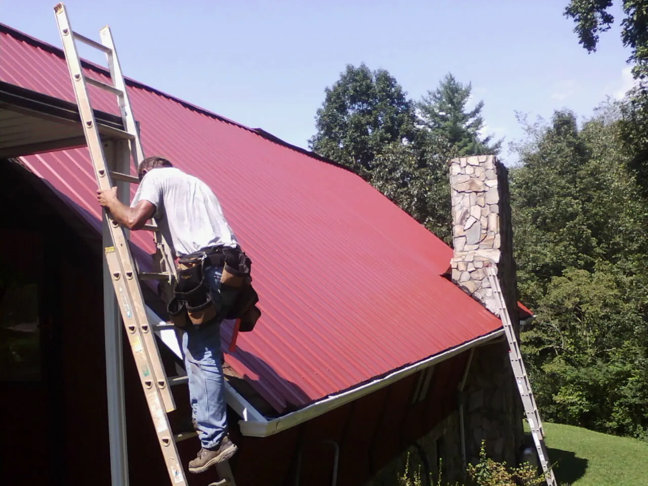 Roofer on a ladder beside a red metal roof and stone chimney — Soffit & Fascia Repair by Roofing Brothers in Branchville